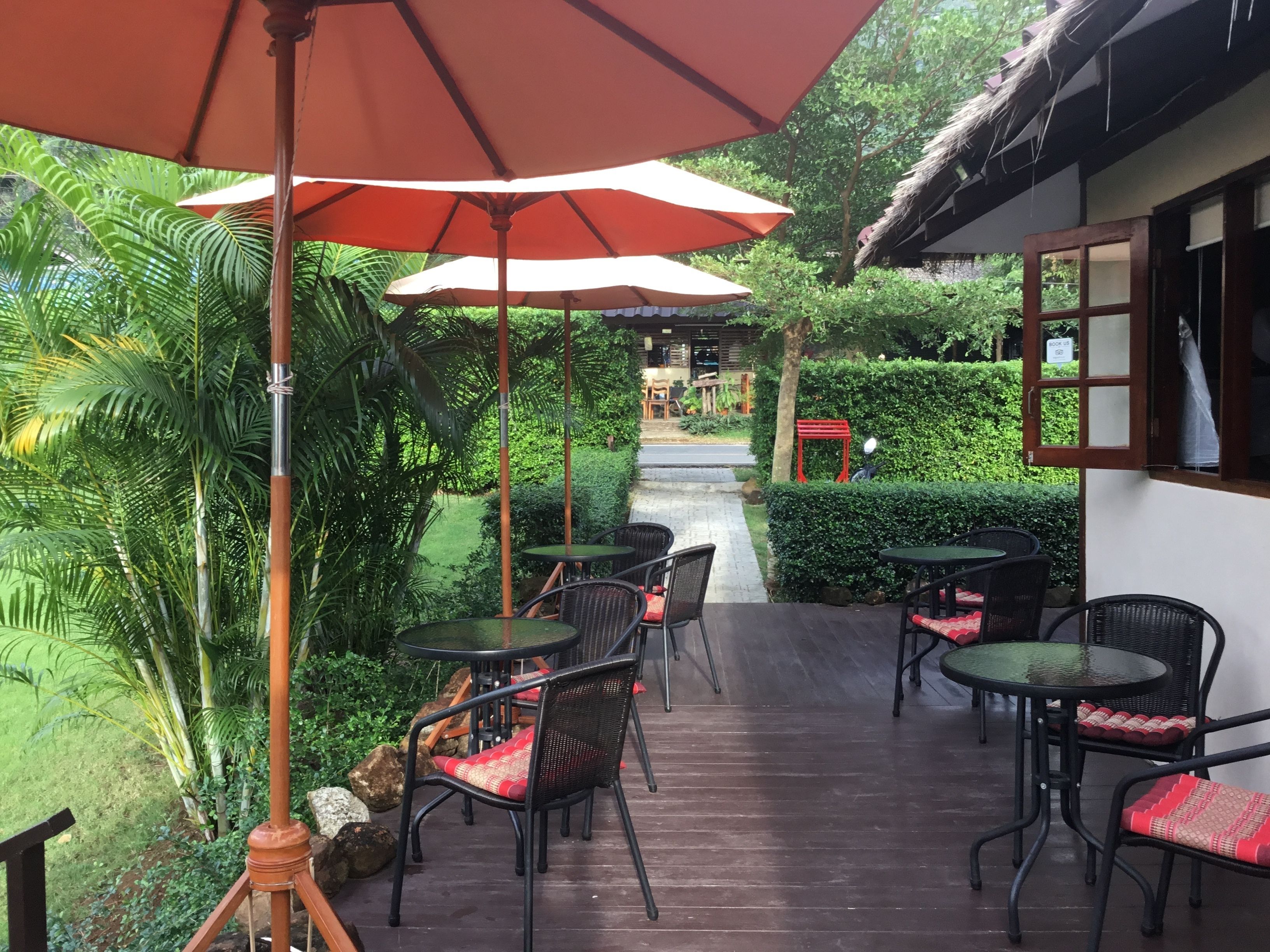 An outdoor patio with tables and chairs, surrounded by lush greenery and a thatched-roof building.