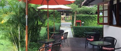 An outdoor patio with tables and chairs, surrounded by lush greenery and a thatched-roof building.