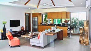 A modern living room with a sofa, a coffee table, and a TV. There is a kitchen area with a refrigerator and a sink. - Ka Villa Rawai : Peaceful 4 Bedrooms (Rawai)