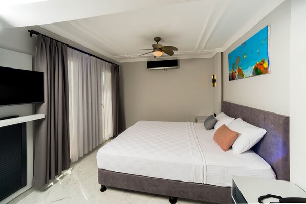 A hotel room with a bed, a ceiling fan, a painting, and a TV.