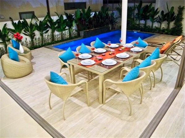 Un comedor con una mesa de vidrio rodeada de sillas de mimbre, una piscina al fondo y plantas tropicales.