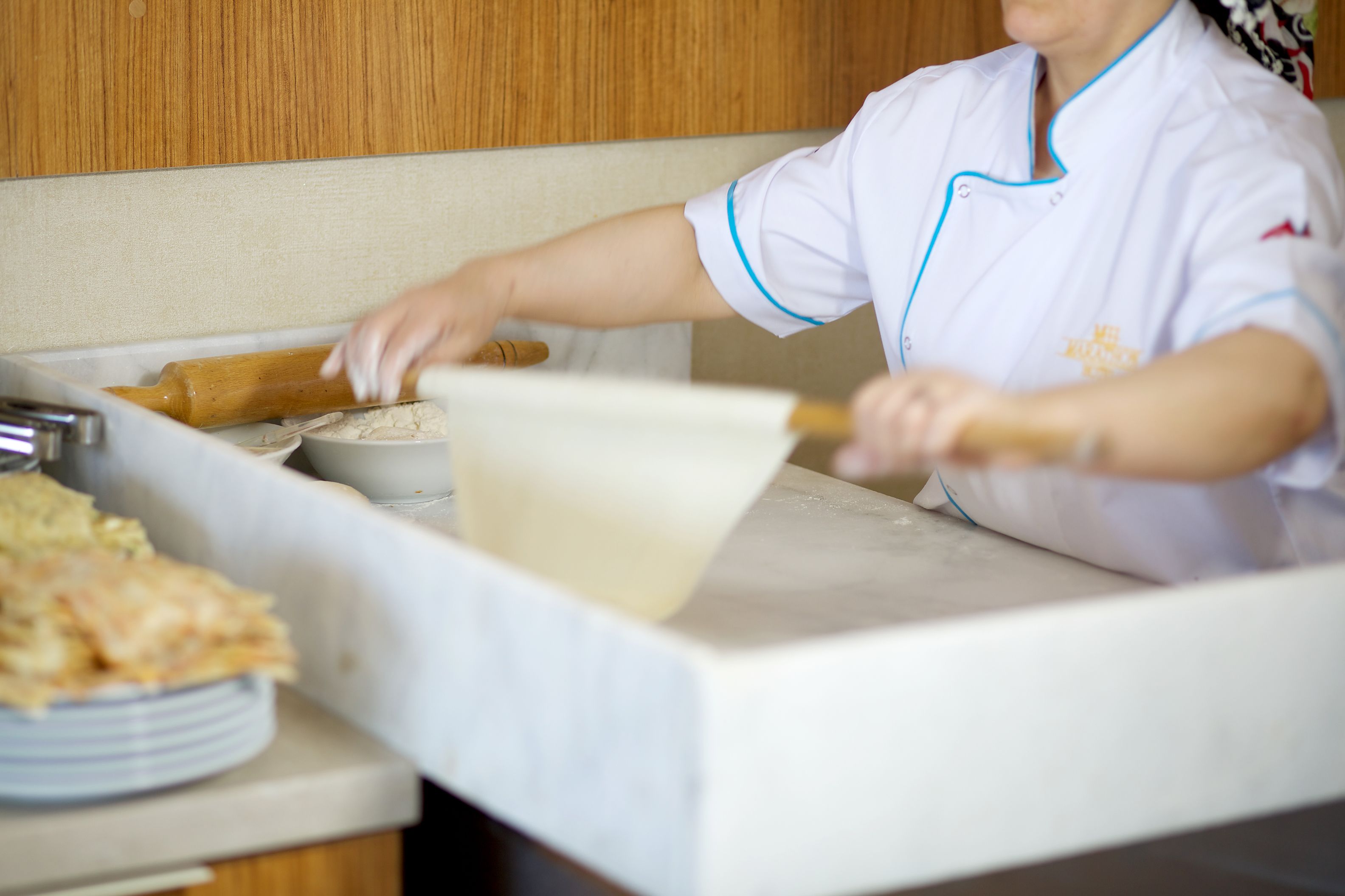 Een persoon in een wit uniform met blauwe accenten die deeg uitrolt op een aanrecht in een keuken.