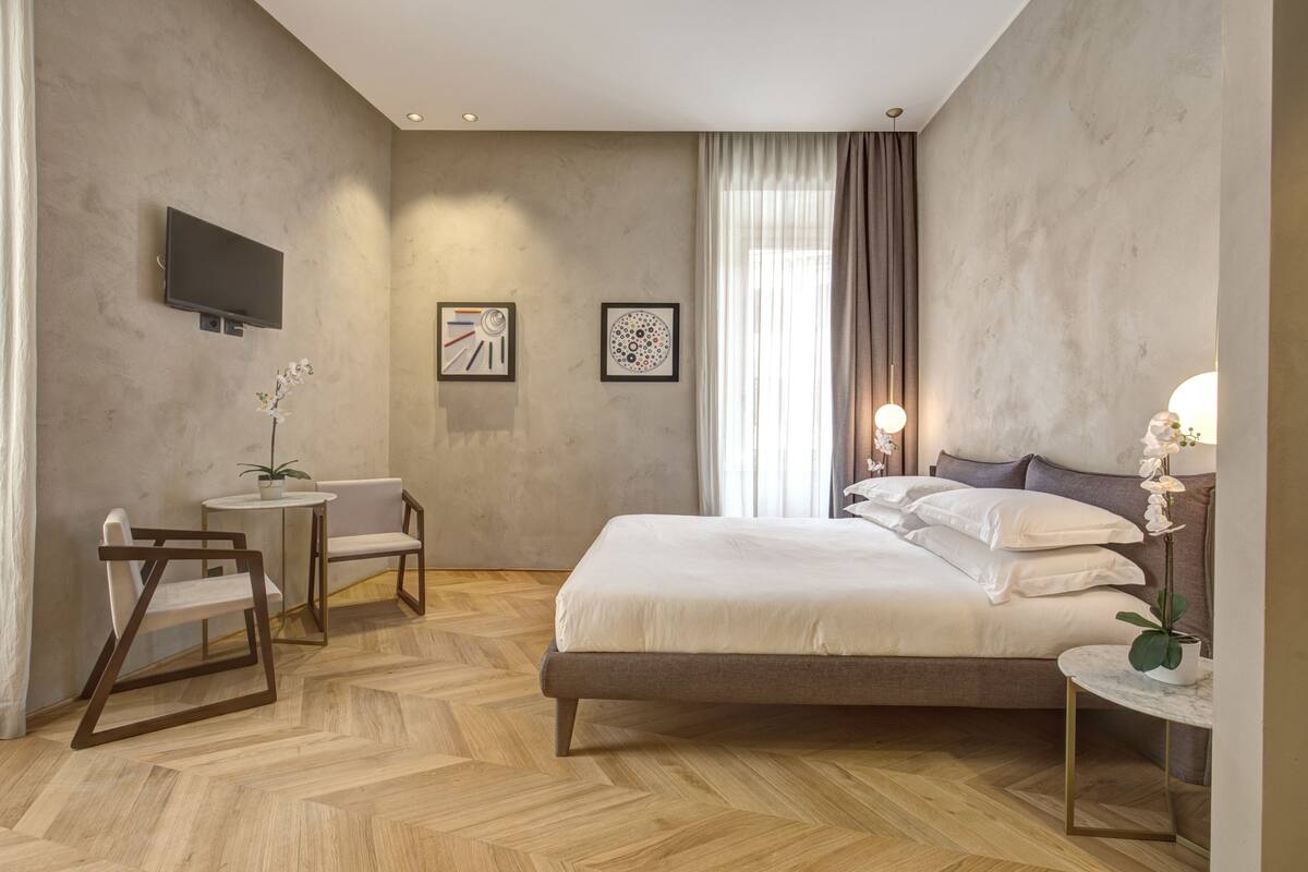 Un dormitorio moderno con una cama grande, un televisor montado en la pared, una mesita con una planta y una silla.