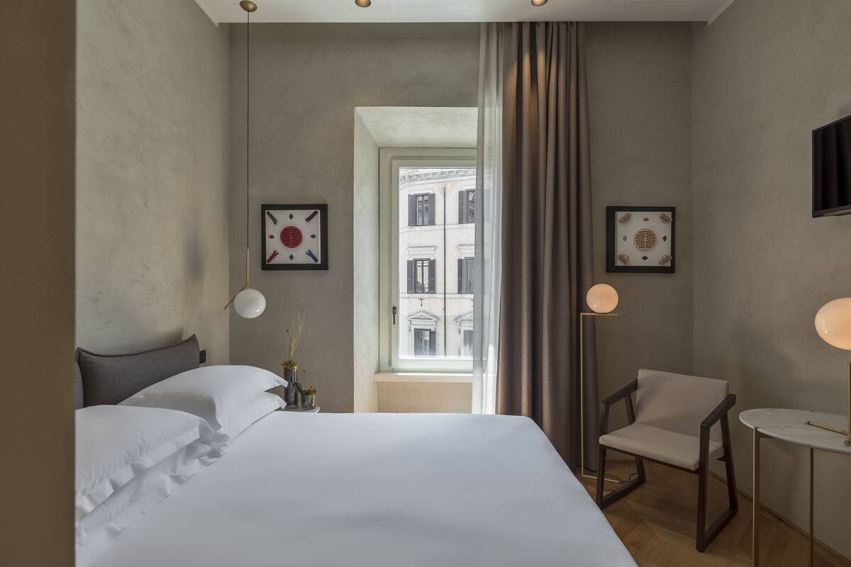 Habitación de hotel con una cama grande, una silla, una mesita y dos lámparas fijadas a la pared. Se ve una ventana con vistas a edificios.