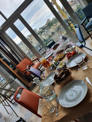Mesa de jantar posta com diversos pratos e bebidas, com vista para edifícios e um parapeito azul ao fundo.