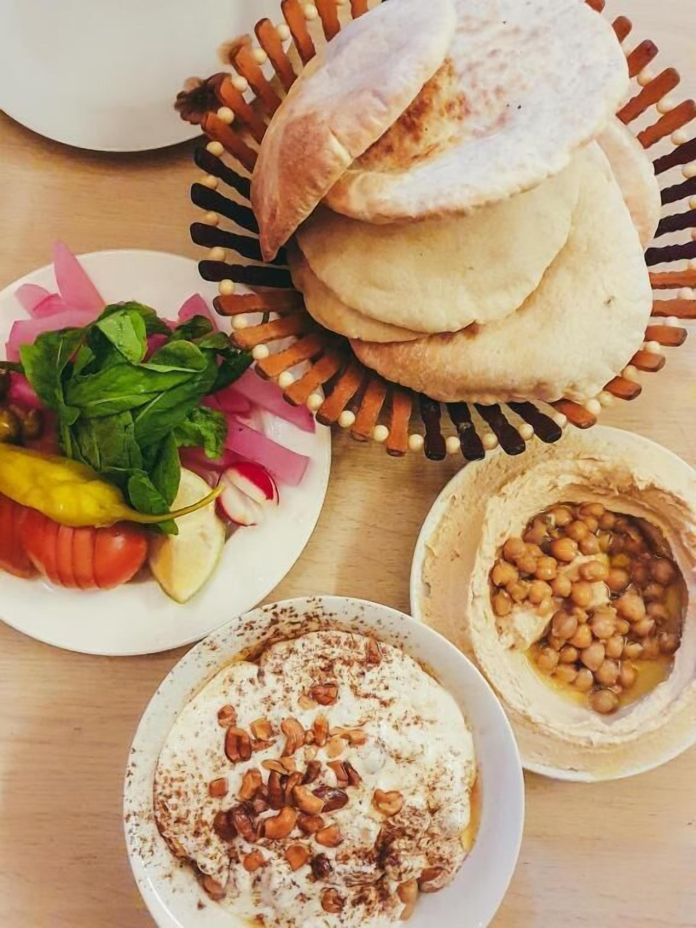 A table with a stack of flatbreads, a bowl of chickpeas, a plate of vegetables, and a bowl of yogurt topped with nuts.