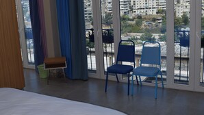 A bedroom with a large bed, a chair, and a view of a cityscape through large windows. - Warda وَرْدَة Lebanese Guesthouse (Zouk Mosbeh)