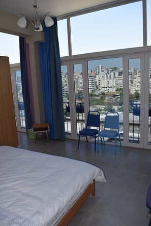 Ein Schlafzimmer mit einem großen Bett, einem Sessel und Blick auf eine Stadtlandschaft durch große Fenster.