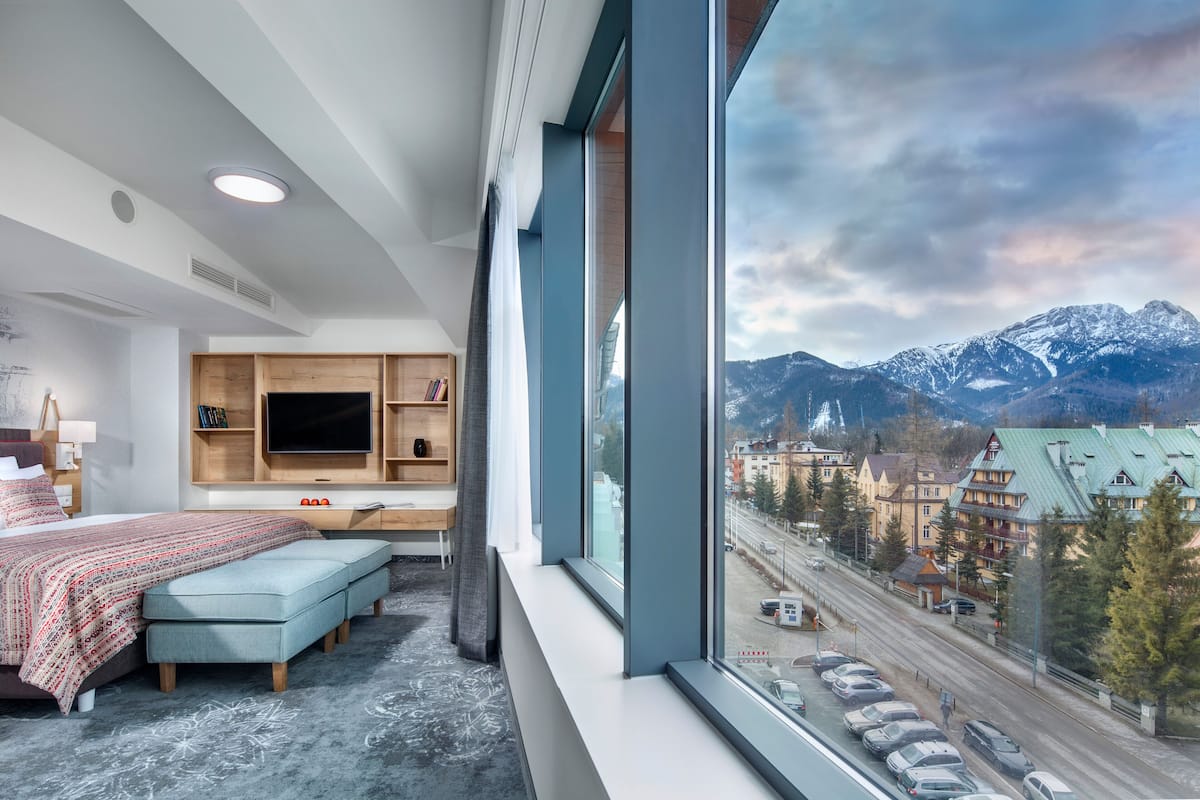 A modern hotel room with a large window, a bed, a desk, and a TV.