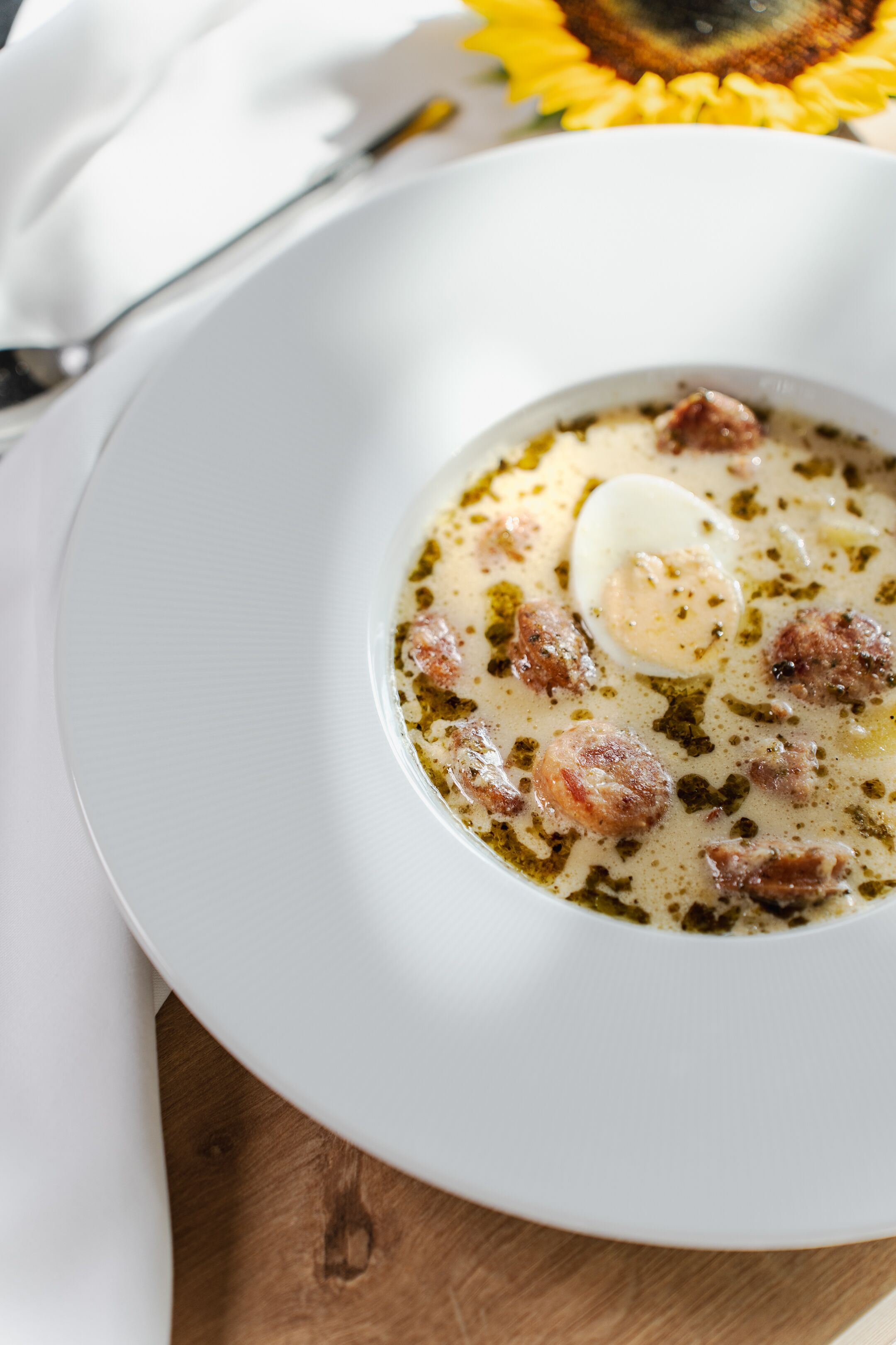 A white plate with a creamy soup containing meat and a poached egg, placed on a wooden surface with a sunflower in the background.