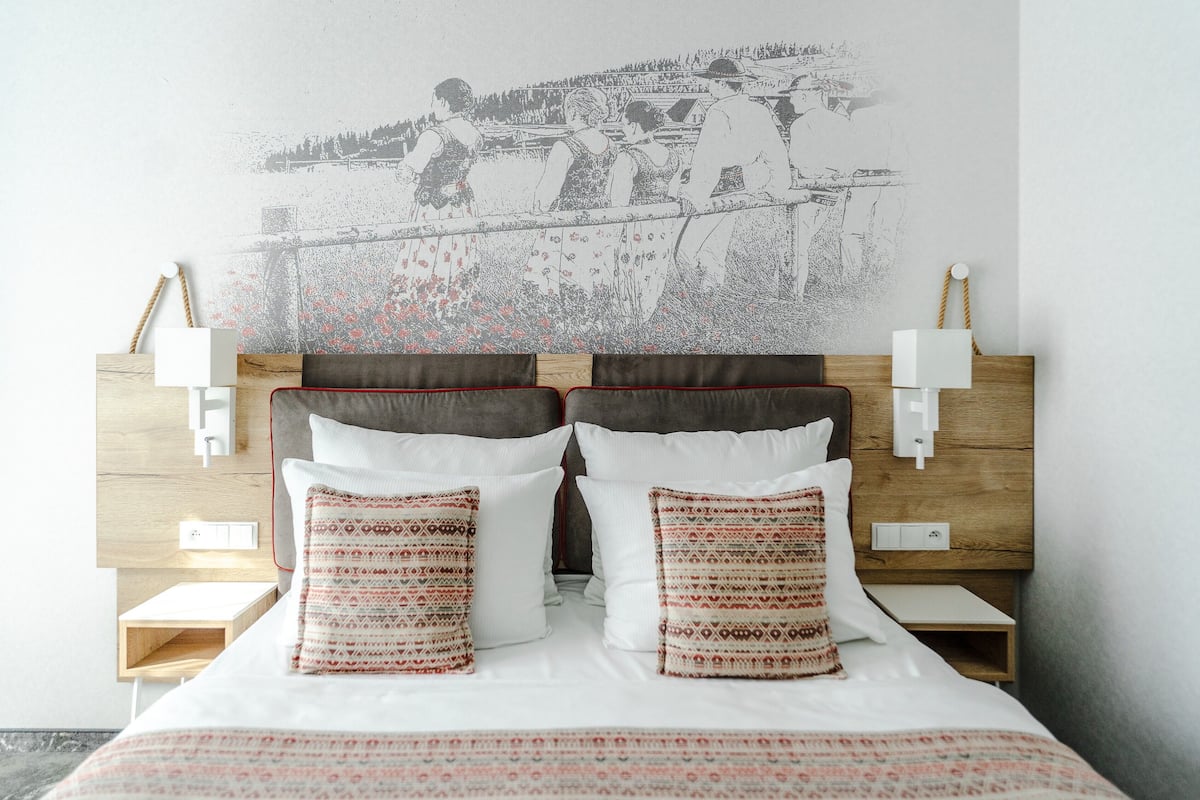A bed with a patterned bedspread, two pillows with geometric designs, a headboard with a wall-mounted lamp, and a wall art depicting a historical scene.
