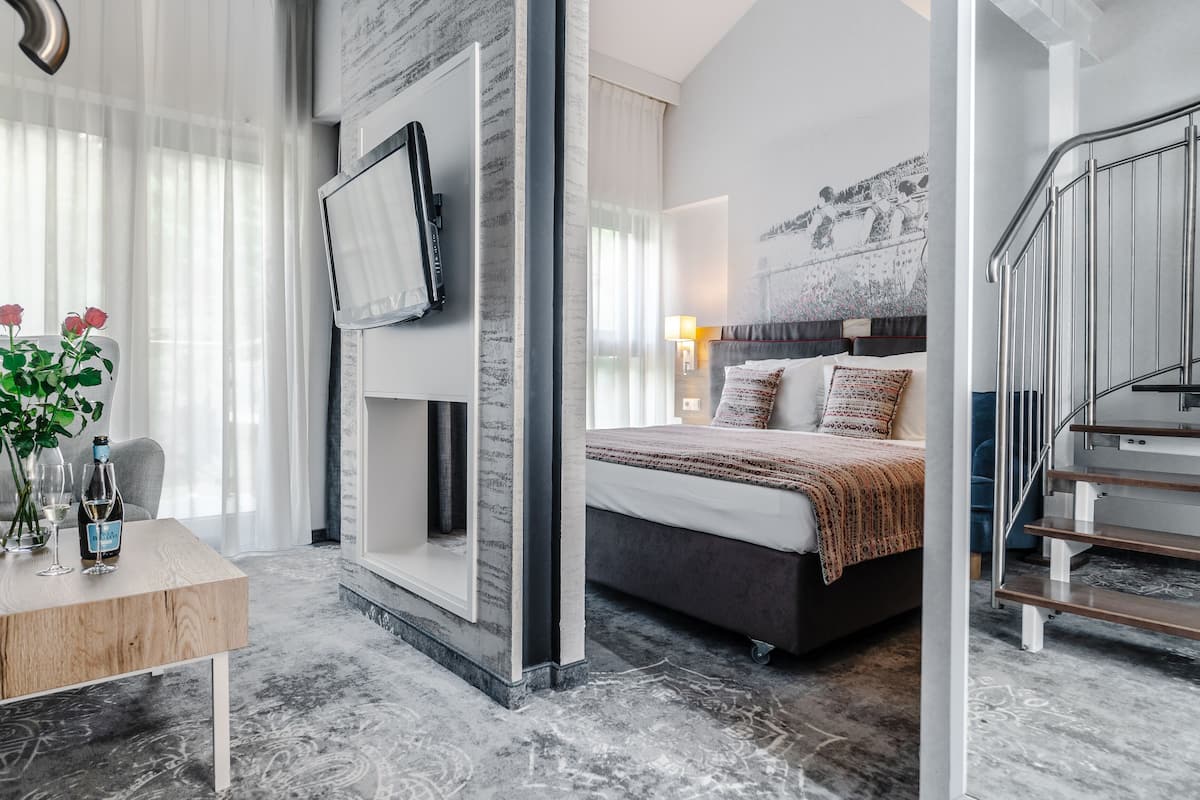 A modern hotel room with a large bed, a flat-screen TV mounted on the wall, a wooden table with a vase of flowers, and a staircase with a metal railing.