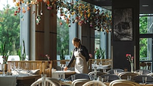 Un restaurant doté de mobilier en bois, de décorations florales suspendues et d’une grande fenêtre offrant une vue sur la verdure.