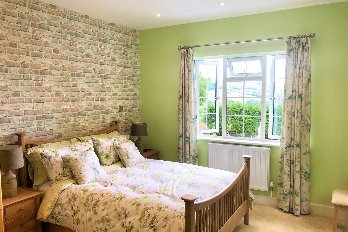 A bedroom with a brick wall, a wooden bed, a nightstand with a lamp, and a window with curtains.