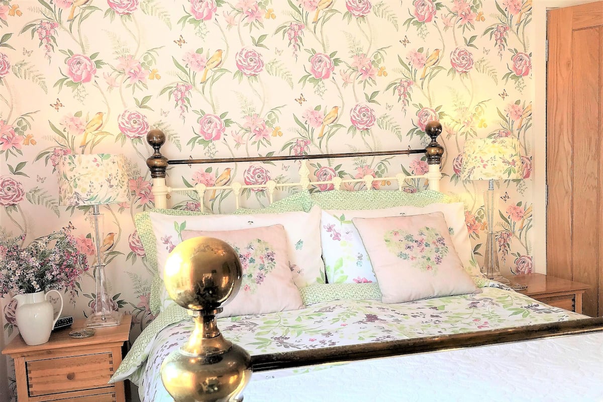 A bedroom with floral wallpaper, a wooden headboard, and a bedside table with a lamp and a vase of flowers.