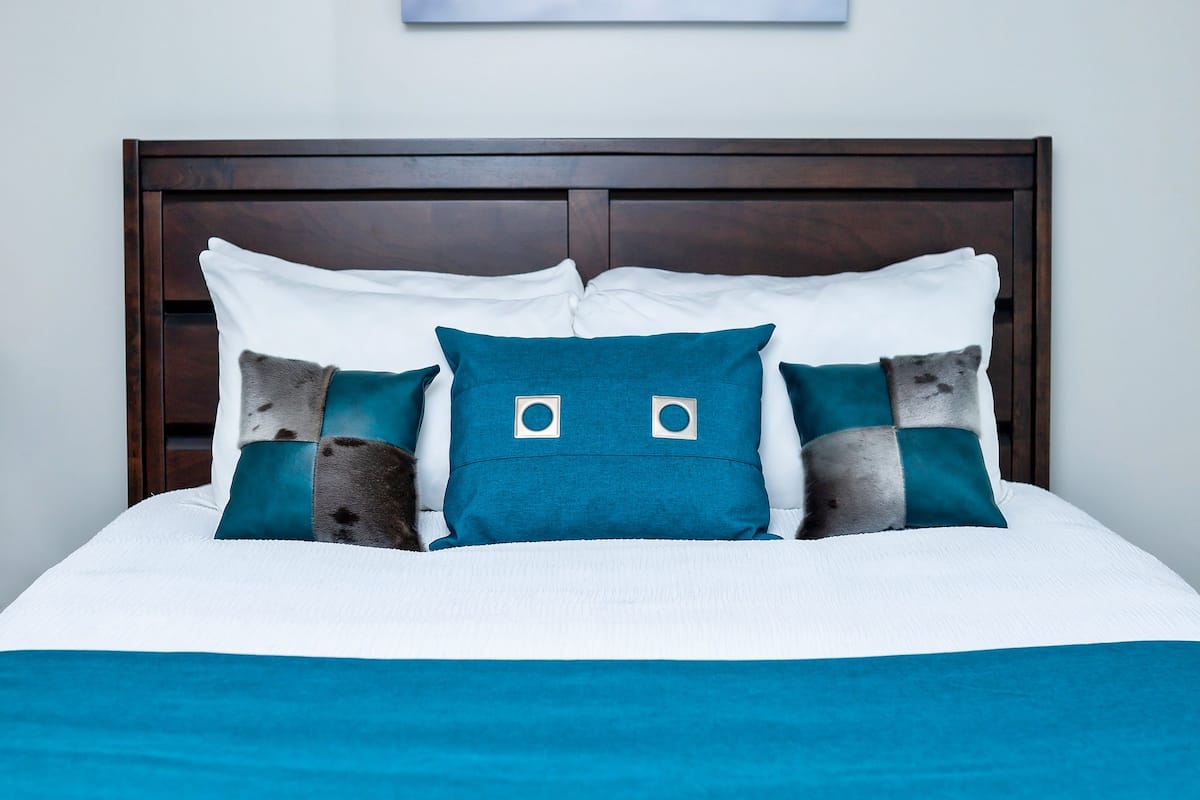 A bed with a white and teal bedspread, a dark wooden headboard, and three teal pillows with a square design.