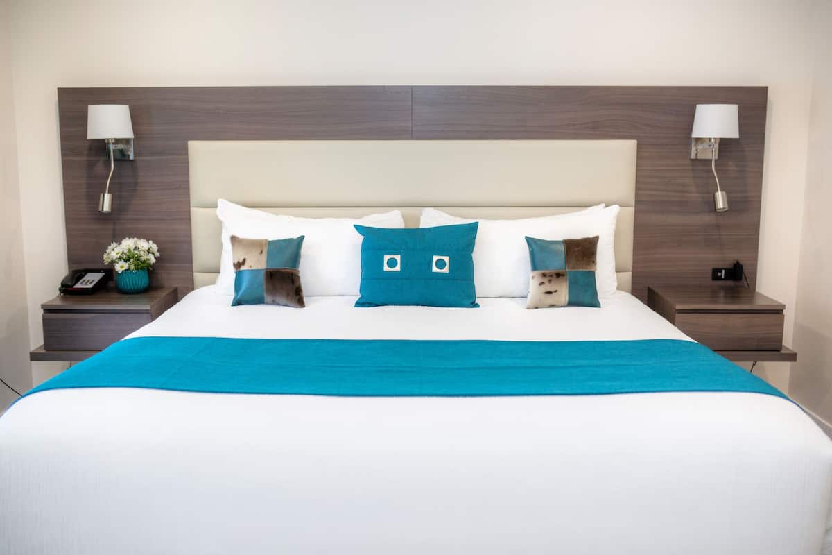 A neatly made bed with a white and blue bedspread, a teal accent pillow, and a matching throw blanket. There are two bedside tables with lamps and a vase of flowers.