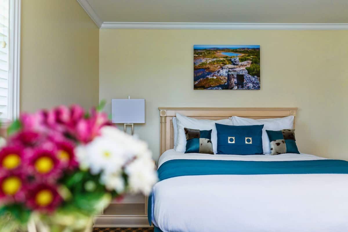 A neatly made bed with a blue and white color scheme, a painting of a landscape on the wall, and a vase of flowers on the nightstand.