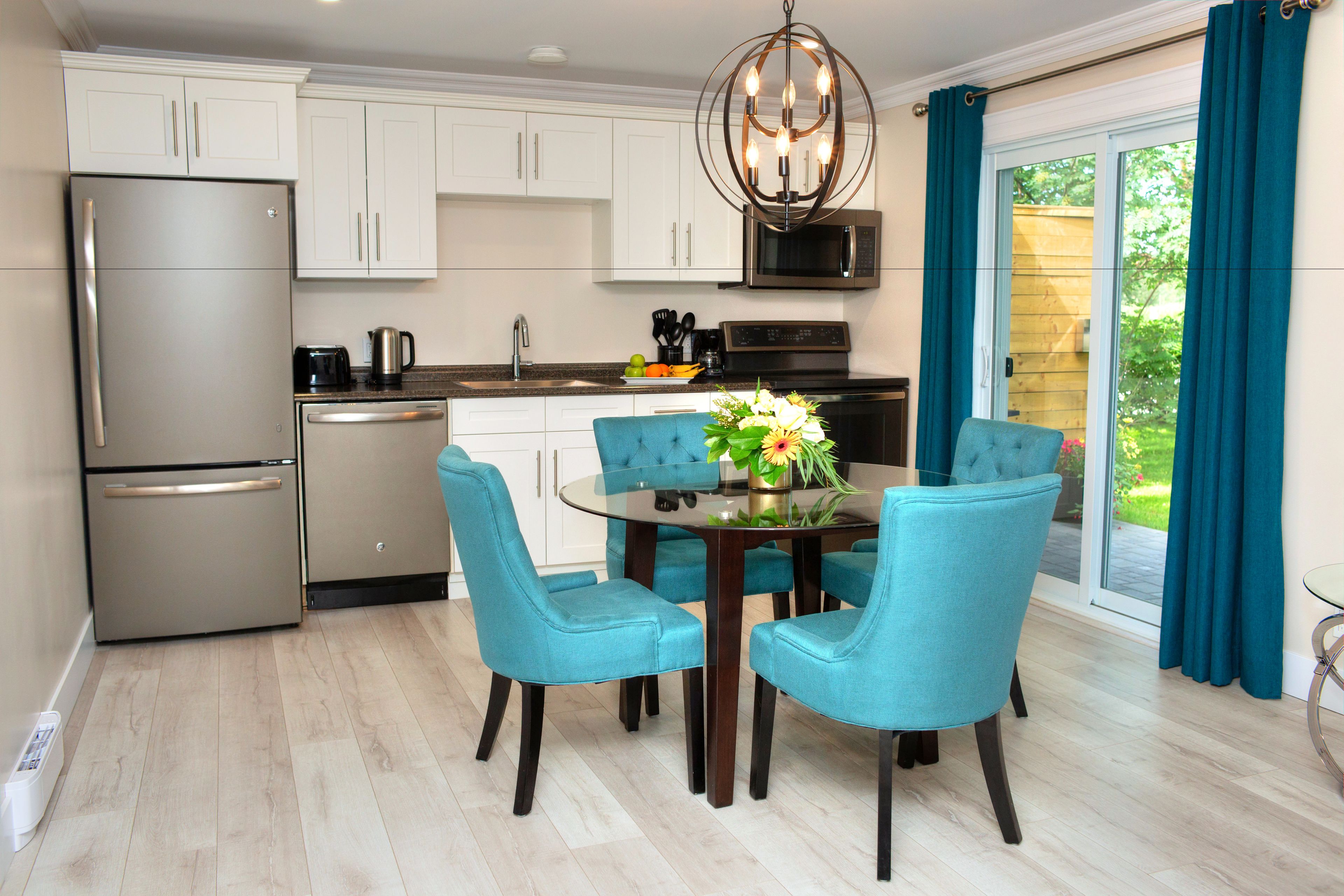 A modern kitchen with white cabinets, a stainless steel refrigerator, and a dining area with teal chairs and a glass table.