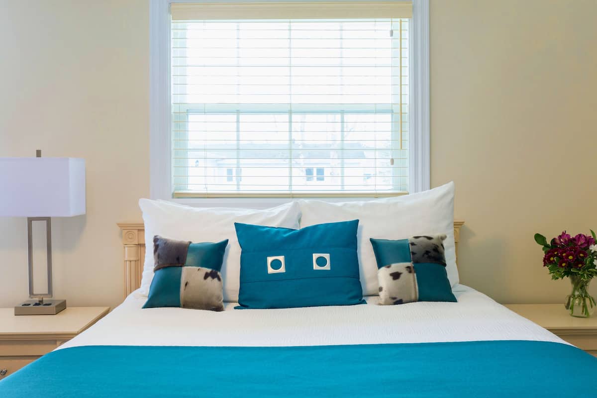 A neatly made bed with a white and teal bedspread, two matching teal pillows, and a white bedside table with a lamp.