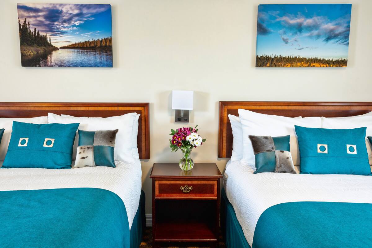 A hotel room with two beds, teal bedding, a nightstand with a lamp, and two landscape paintings on the walls.