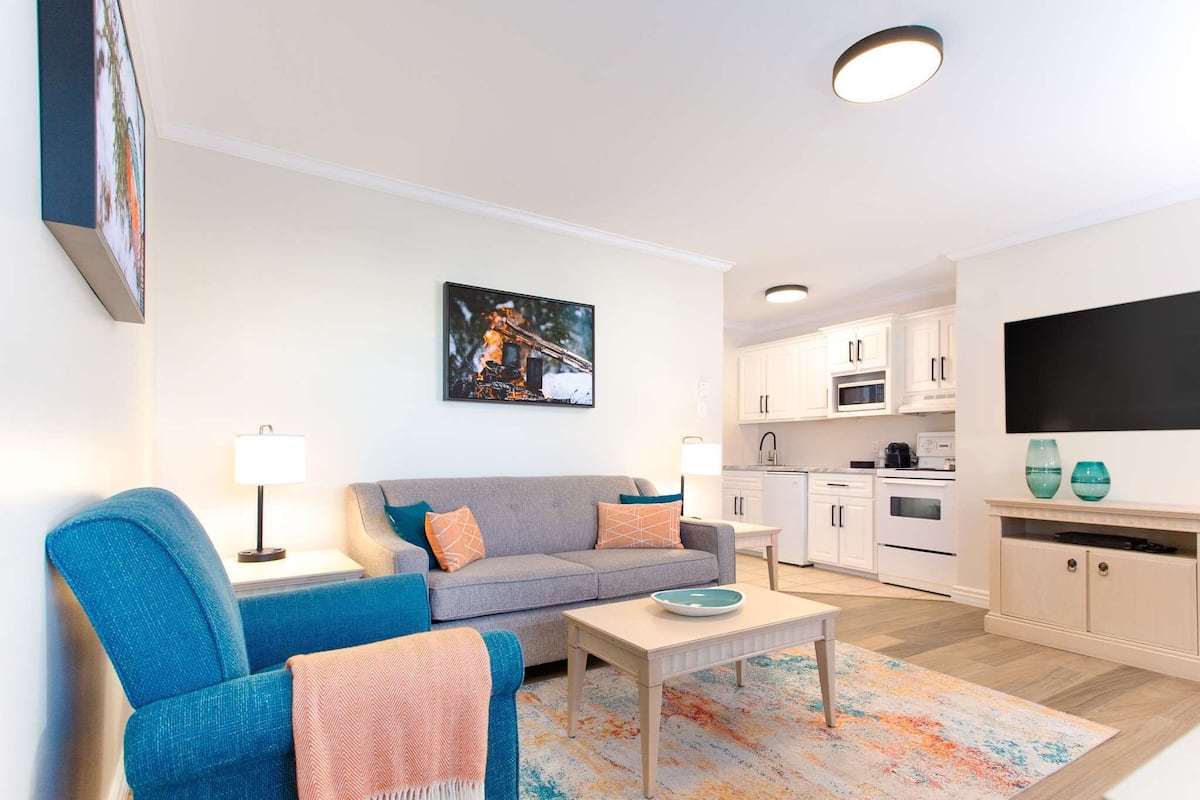 A modern living room with a sofa, armchair, and coffee table, a kitchen area with cabinets and appliances, and a flat-screen TV mounted on the wall.