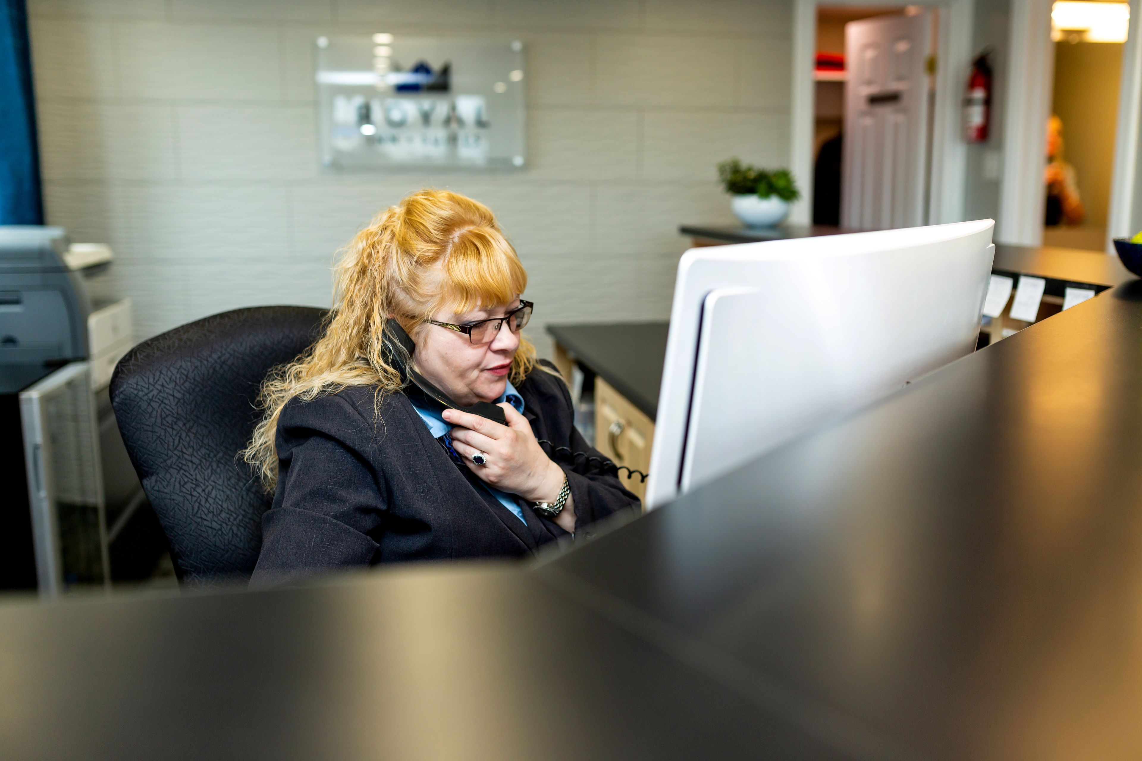 Una persona sentada en un escritorio con una computadora, en una habitación con pared de azulejos y una puerta al fondo.