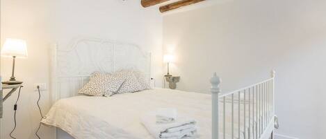 A bedroom with a white bed, two bedside lamps, a white headboard, and exposed wooden beams on the ceiling.