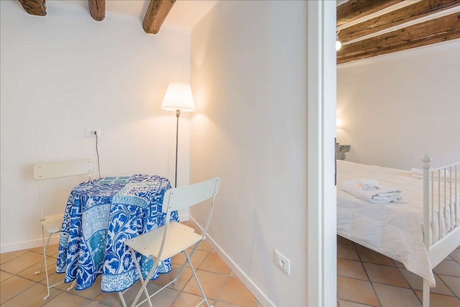 Une pièce comprenant un lit, une table recouverte d’une nappe à motifs bleus, une chaise et un lampadaire.