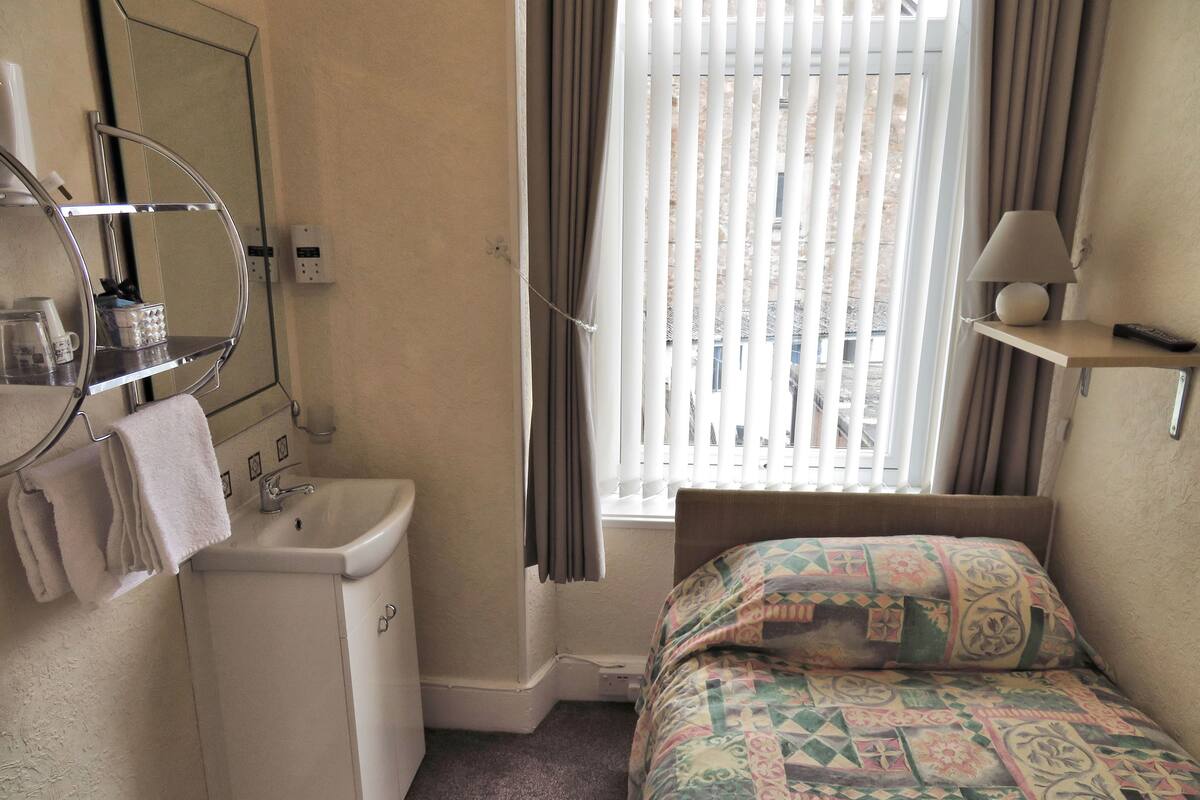 Ein kleines, helles Schlafzimmer mit einem Einzelbett, einem Nachttisch, einem Spiegel und einem Fenster mit Jalousien.