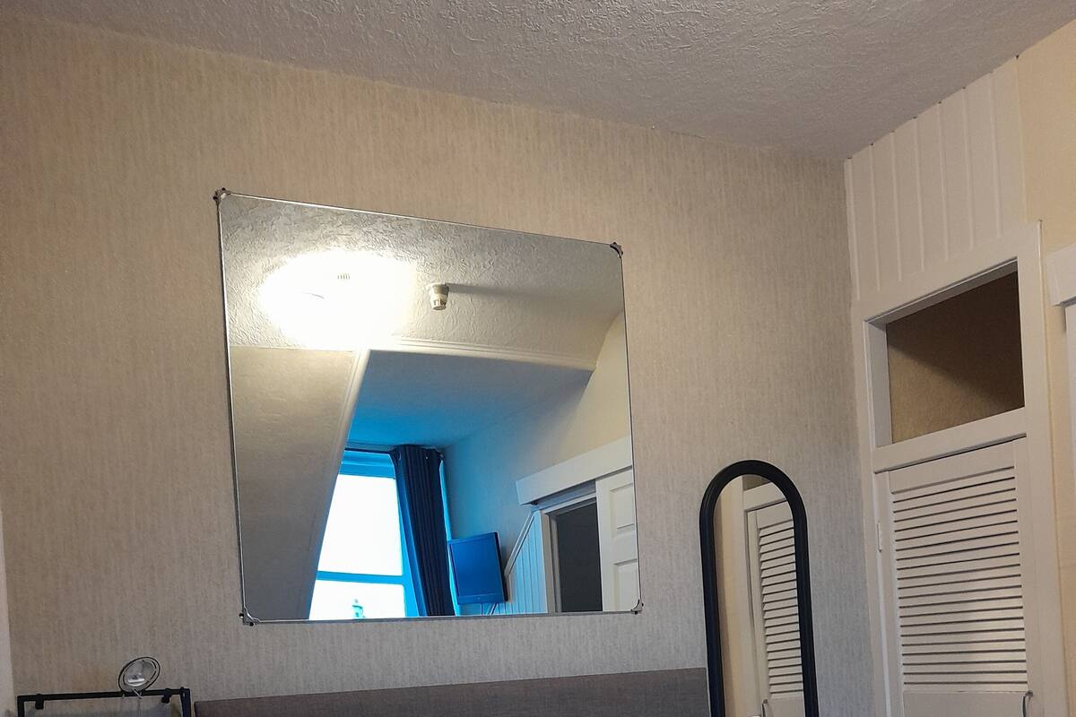 Ein ordentlich bezogenes Bett mit einer braunen Steudecke, ein Nachttisch mit einer Lampe, ein Spiegel an der Wand und ein Fenster mit Vorhängen.
