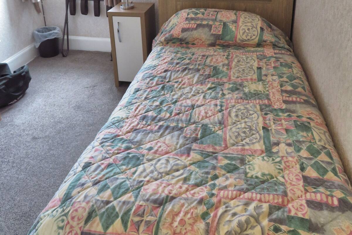 Ein kleines Zimmer mit einem Einzelbett, einem Holzkopfende, einem Nachttisch mit Lampe und einem Fenster mit Vorhängen.