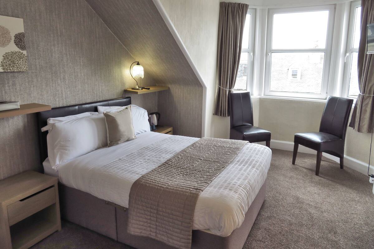 Ein Hotelzimmer mit Bett, Nachttisch, Stuhl, Schreibtisch und einem Fenster mit Vorhängen.