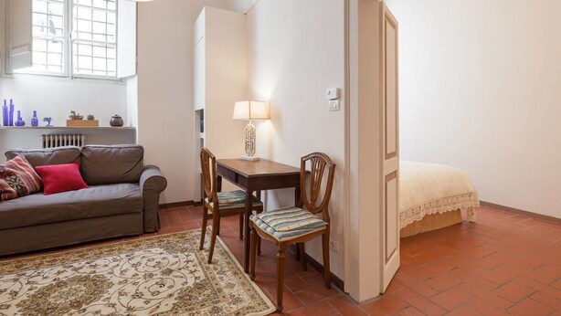 A cozy room with a brown leather sofa, a wooden dining table with chairs, a patterned rug, a bed with a white duvet, and a window with blinds. - Suite Elda (Verona)