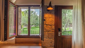 A room with a stone wall, a wooden door, and a large window with a view of a garden. - Assos Park Hotel (Ayvacik)