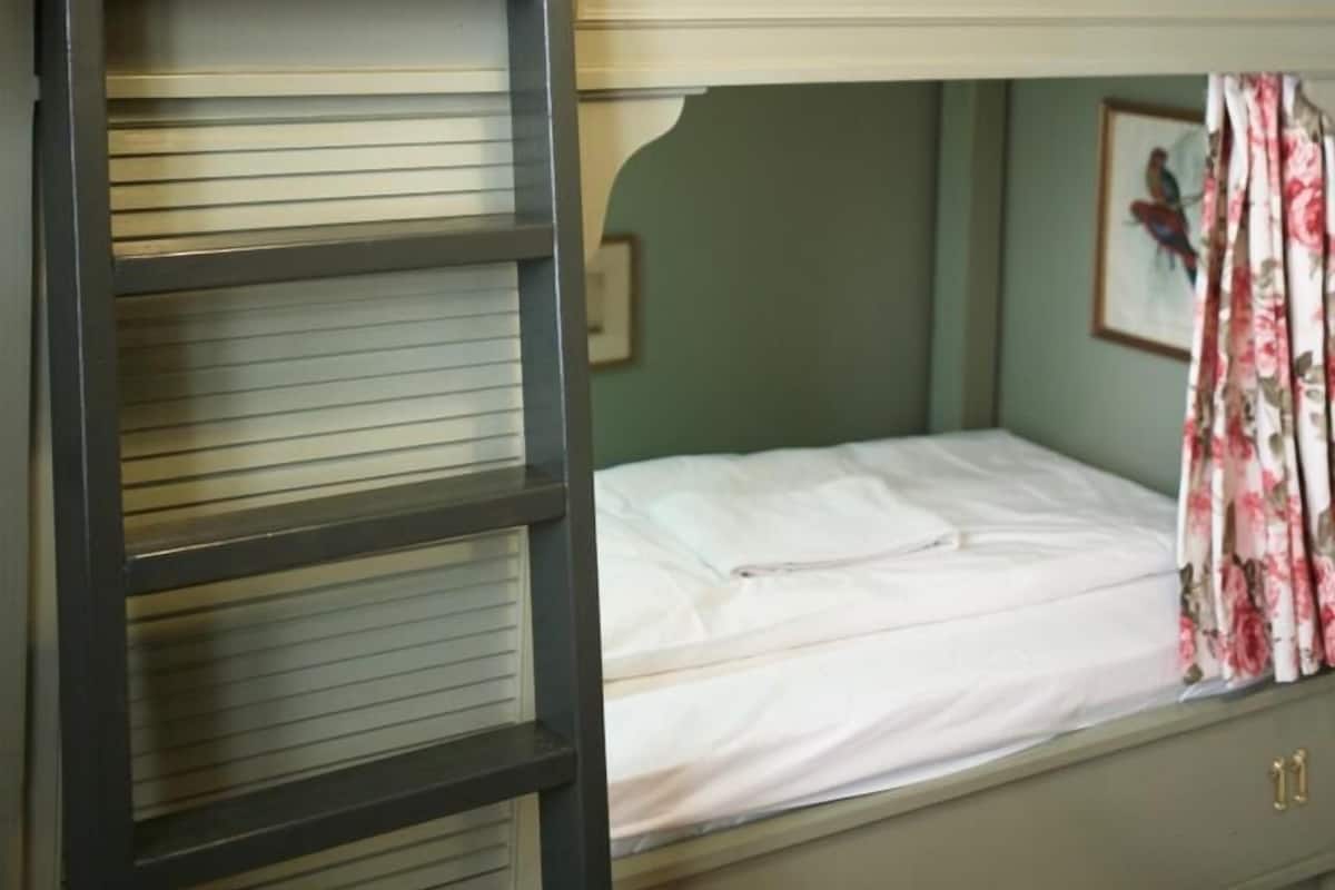 A bunk bed with a ladder, a single bed with a floral curtain, and a wall-mounted light switch.