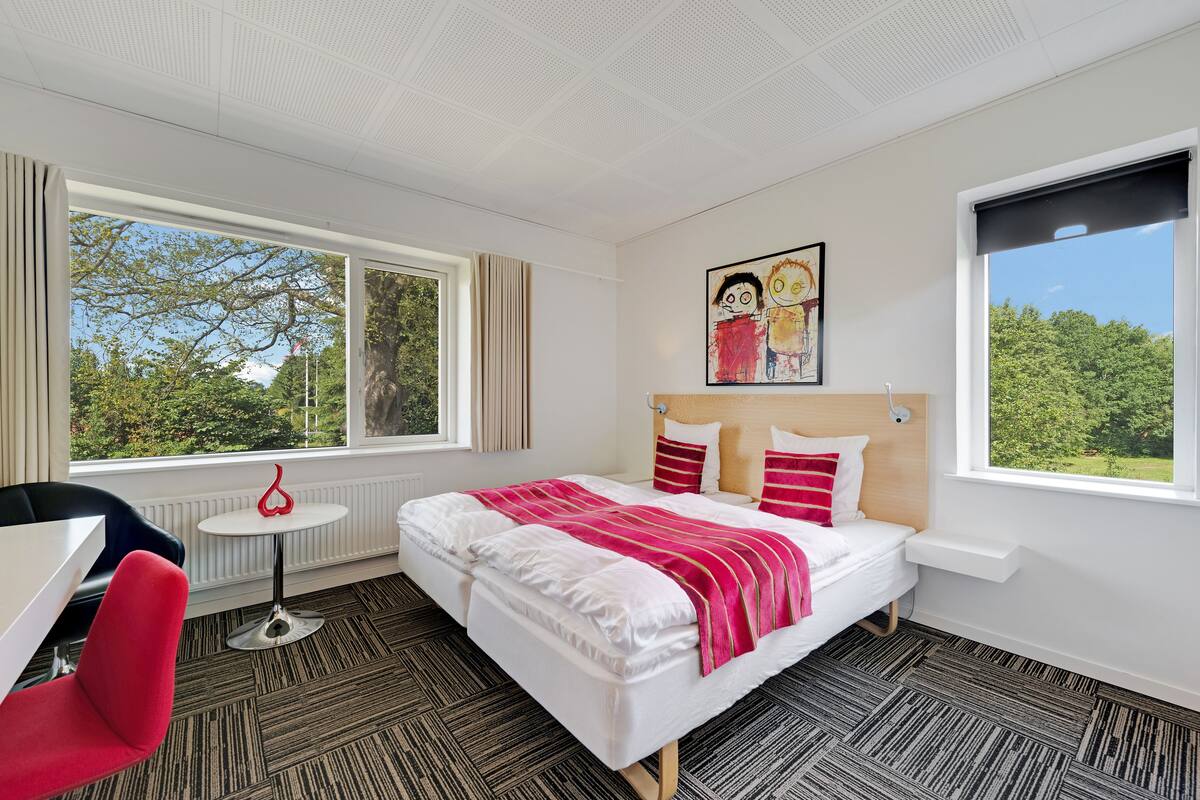 Une chambre d’hôtel avec un grand lit, une petite table, une chaise et deux fenêtres donnant sur la verdure.