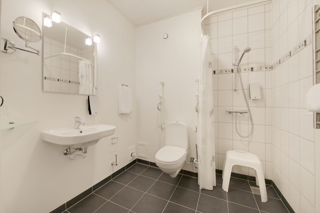 A modern bathroom with a white sink, mirror, toilet, shower, and a small white stool.