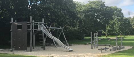 A playground with a slide and climbing structures, surrounded by trees and grassy areas.