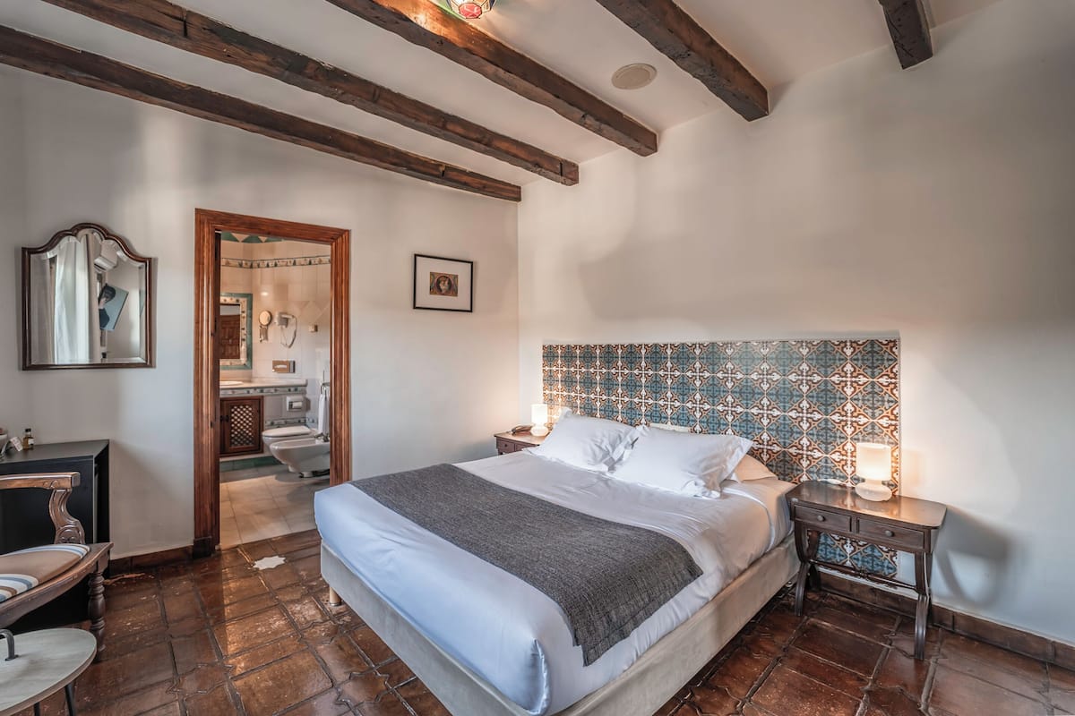 Ein Schlafzimmer mit einem großen Bett, Holzbalken, einem gemusterten Kopfteil, einem Nachttisch mit Lampe und einem durchgehenden Türspalt erkennbaren Badezimmer.