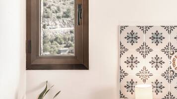 A room with a window overlooking a mountainous landscape, a potted plant, and a lamp on a wooden stool.