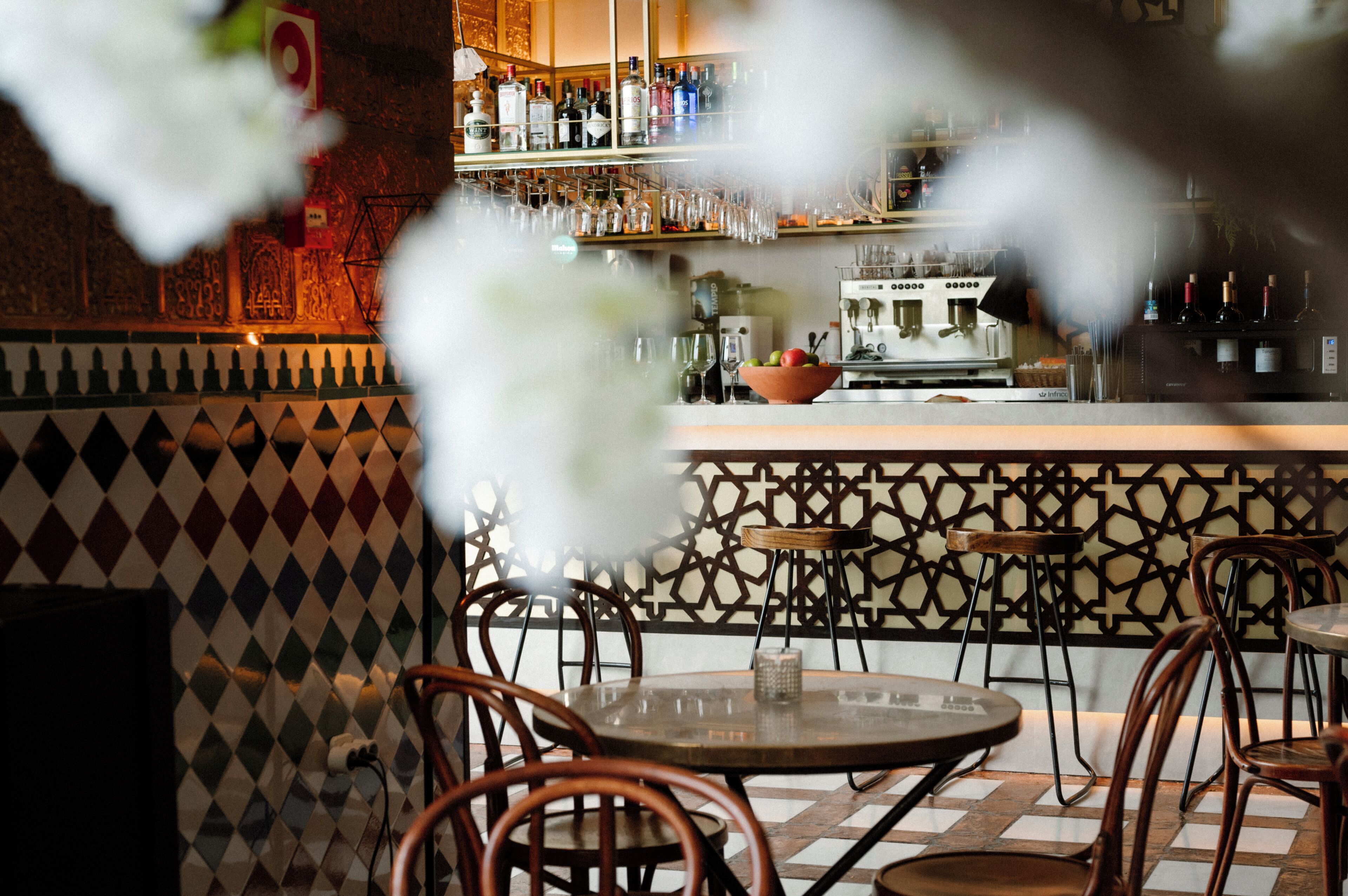 A bar area with a patterned wall, a counter with bottles and glasses, and round tables with chairs.