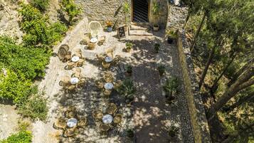 An outdoor seating area with wicker tables and chairs, surrounded by potted plants and a stone wall.