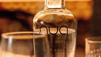 Une bouteille en verre transparent, munie d’un bouchon noir portant l’étiquette « Aqua », est posée sur une table, accompagnée de deux verres, dont l’un contient une tranche de citron.