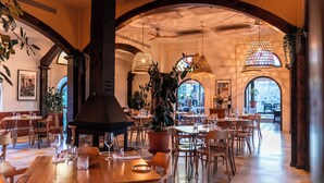 Een restaurant met houten tafels, stoelen en een piano. Er zijn hangende lampen en potplanten.