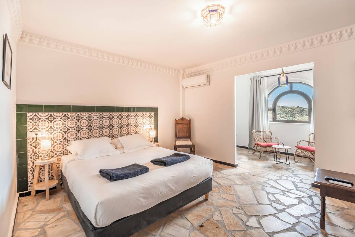 Ein Schlafzimmer mit einem großen Bett, einem Nachttisch, einem Sessel, einem Tisch und Blick ins Freie durch ein Fenster.