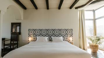 Une chambre à coucher avec un grand lit, une chaise en bois, une tête de lit à motifs et une fenêtre avec des rideaux.
