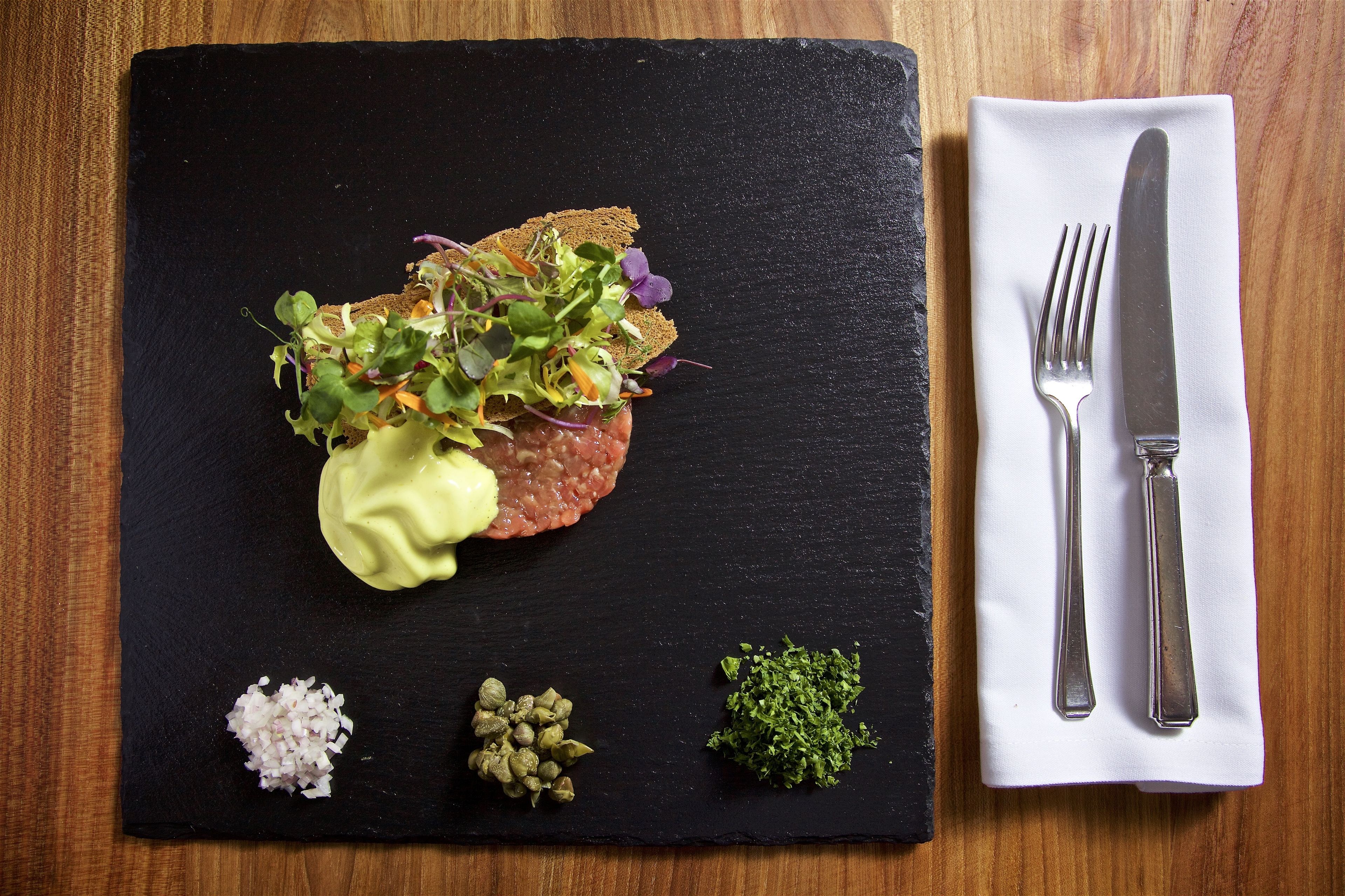 A black slate serving board with a meat patty topped with greens, a dollop of sauce, and garnishes of chopped onions, capers, and herbs.