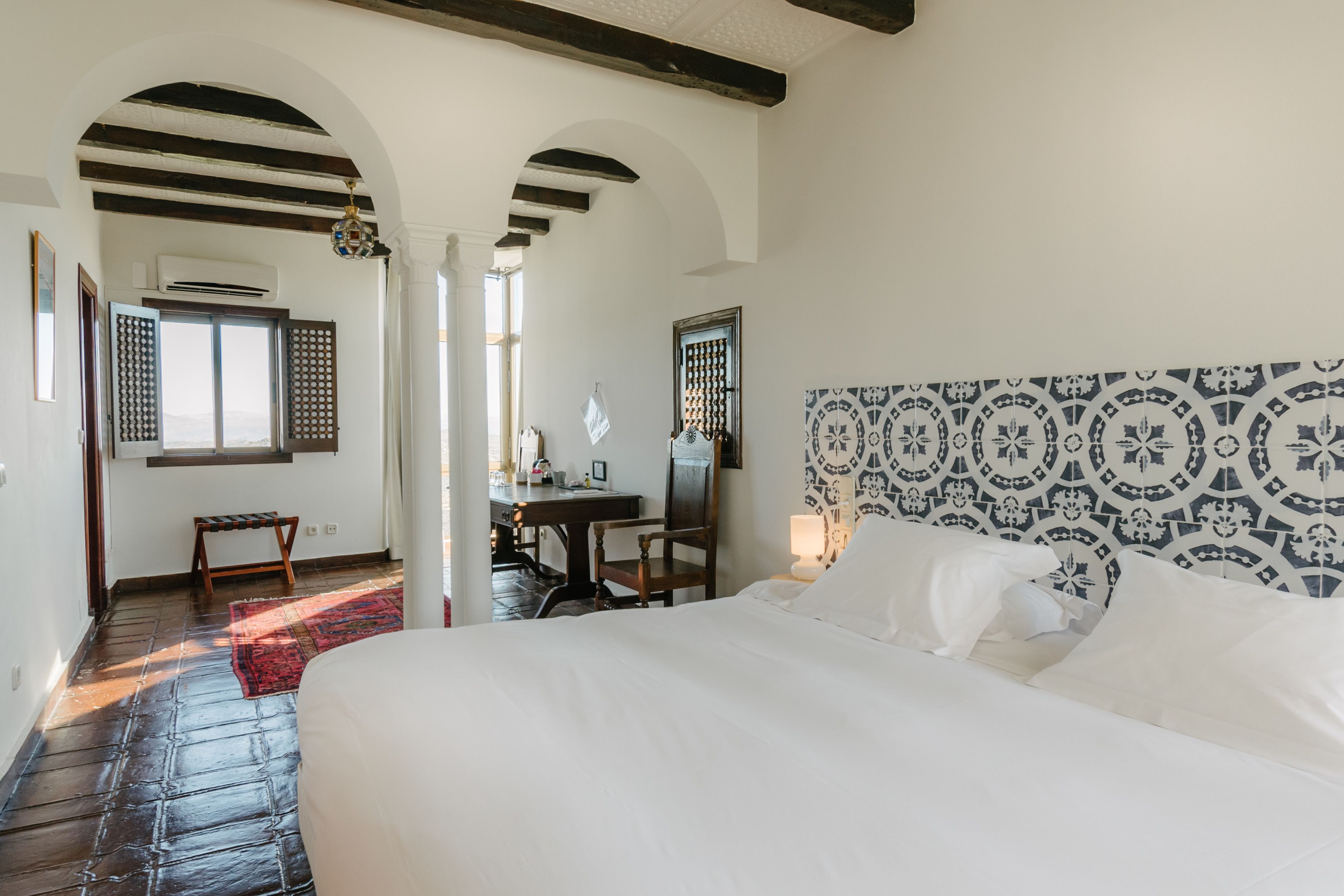 A hotel room with a large bed, a desk, and a chair. There is a window with shutters and a view of the outside.