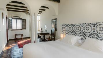 A hotel room with a large bed, a desk, and a chair. There is a window with shutters and a view of the outside.